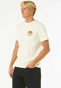 Cremfarbener Baumwoll-T-Shirt mit kurzen Ärmeln. Verfügt über eine Retro-Regenbogen-Grafik und Text im Bereich der linken Brust. Kombination mit schwarzen Jeans.