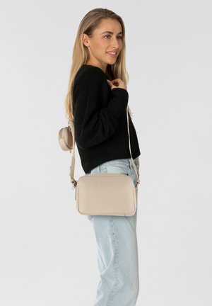 Borsa a tracolla in pelle beige con logo impresso, forma rettangolare, chiusura con zip e tracolla regolabile, abbinata a un maglione nero e jeans chiari.