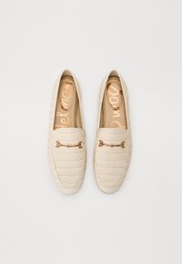 Beige loafers ar krokodila rakstu, ar zelta detaļu akcentu un mīkstu zoli, kurā ir zīmola nosaukums zeltā.