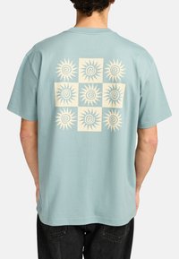 Hellblaues Baumwoll-T-Shirt mit einem großen cremefarbenen Sonnengrafikmuster auf der Rückseite, das neun spiralförmige Sonnendesigns in quadratischen Sektionen zeigt.