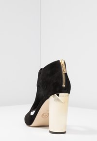 Musta samettinen korkokenkä, jossa on kultasävyinen uritettu korko ja takana vetoketjusuljenta. Vetojaulasta löytyy avonainen varvas ja leikattu muotoilu sivuilla.