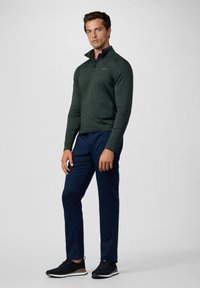 Pulover de manga larga verde oscuro con cuello alto, combinado con pantalones azul marino y zapatillas negras, de pie sobre un fondo neutro.