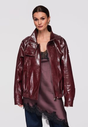 Kvinde iført en glinsende bordeauxrød læderjakke over en satin natkjole med sort blonderkant, kombineret med mørkeblå jeans, foran en ensartet baggrund.