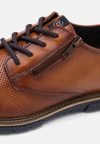 Chaussure pour homme en cuir marron avec détails perforés, lacets bleu marine, fermeture éclair latérale et semelle en caoutchouc noire.