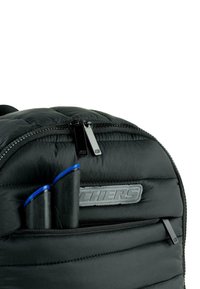 Mochila negra acolchada con una textura suave, bolsillo con cremallera y dos bolígrafos con puntas azules visibles en el bolsillo delantero.