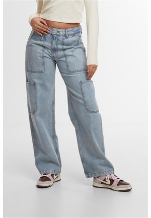 Personne portant un jean large bleu clair avec de grandes poches plaquées et des baskets rose-brun, main posée sur la hanche.