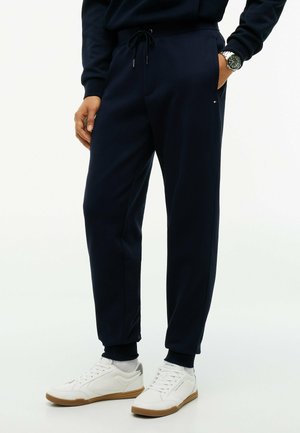 Homme portant un pantalon jogger bleu marine avec une taille à cordon, des baskets blanches et une montre-bracelet, debout avec une main dans la poche.