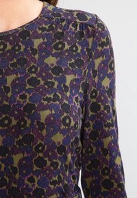 Długi rękaw bluzka w oliwkowej zieleni z kwiecistym wzorem w kolorze fioletowym i czarnym. Okrągły dekolt z dyskretnymi przeszyciami na ramionach.