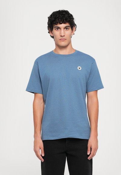 Blaues Baumwoll-T-Shirt mit kurzen Ärmeln, rundem Ausschnitt und einem kleinen weißen Logo-Patch auf der linken Brust. Getragen mit schwarzen Jeans.