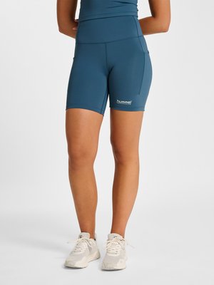 Persoon die blauwe aansluitende sportshorts met zijzakken en witte hardloopschoenen draagt, staand tegen een effen lichte achtergrond.