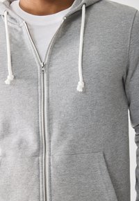 Nahaufnahme einer Person, die einen grauen Reißverschluss-Hoodie mit weißen Kordeln über einem weißen T-Shirt trägt.