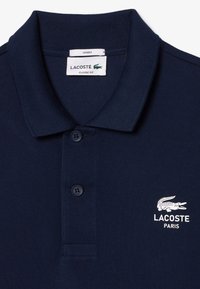 Granatowa koszulka polo z textured materiału, z klasycznym kołnierzykiem, dwoma guzikami oraz białym haftowanym logo i napisem Lacoste.