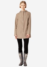 Chaqueta impermeable beige con capucha, con toggles y botones plateados, combinada con botas de lluvia azules con diseño y leggings negros.