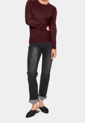 Person iført en vinrød trøje med lange ærmer, sorte cuffede jeans og sorte slip-on sko med sløjfedetalje på en lys baggrund.