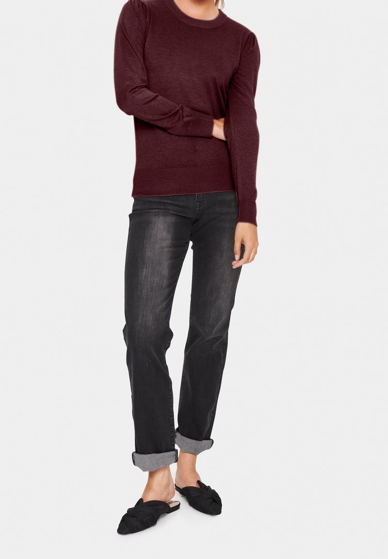 Personne portant un pull bordeaux à manches longues, un jean noir avec revers et des chaussures noires sans lacets avec un détail nœud sur un fond clair.