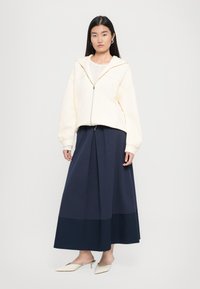 Hnedá mikina na zips s voľným strihom, kombinovaná s tmavomodrou plisovanou maxi sukňou, ktorá má kontrastné panely. Biely podpätkový model. 