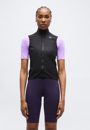 Vrouw met gevlochten haar, gekleed in een zwart mouwloos vest met rits over een paarse wielertenue en donkerblauwe wielershorts, rechtopstaand.