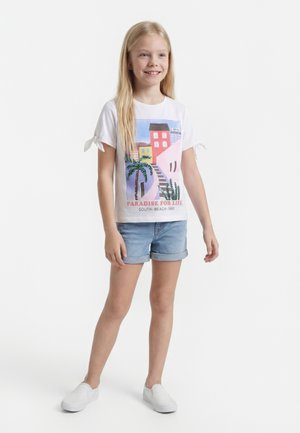 Lachend meisje staand, draagt een wit T-shirt met kleurrijke strandscène print en de tekst "Paradise for Life", denim shorts en witte instapschoenen.