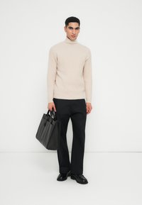 Mann in beiger Rollkragenpullover, schwarzen Hosen und schwarzen Schuhen, der eine große schwarze Ledertasche hält und vor einem weißen Hintergrund steht.