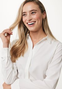 Blusa bianca a maniche lunghe con colletto classico e polsini con bottone, caratterizzata da una leggera texture e dettagli sulla patta frontale.