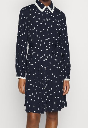 Marineblauwe jurk met lange mouwen, witte polkadots, witte kraag en manchetten, knoopsluiting vooraan en gestrikte taille riem.