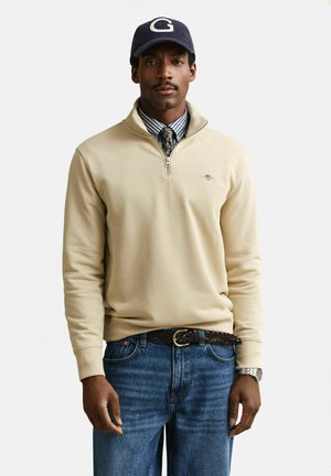 Mann trägt einen beigefarbenen Pullover mit einem Viertelreißverschluss, ein gestreiftes Hemd mit gemustertem Krawatte, blaue Jeans, geflochtener Gürtel, navyblauen Mütze mit "G" und Armbanduhr.