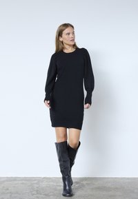 Musta neulemekko pitkilla hihoilla, ribbauslahkeilla ja helman. Yhdistetty polvipituisia mustia nahkasaappaita. Yksinkertainen rakenne, minimalistinen muotoilu.