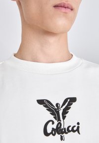 Hvit hettegenser med ribbestrikket hals; har et svart grafikk av en bevinget figur og "Colucci"-tekst i et stilisert skrifttype.