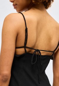 Γυναίκα με μαύρο φόρεμα με λεπτές τιράντες και δέσιμο στην πλάτη, που δείχνει το πάνω μέρος της πλάτης και το αριστερό χέρι σε απλό φόντο.