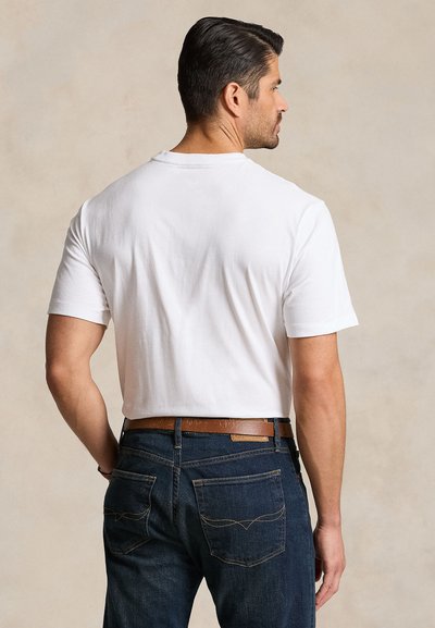 Mand med mørkt hår står med ryggen mod os iført en ensfarvet hvid kortærmet t-shirt, mørkeblå jeans og et brunt læderbælte.