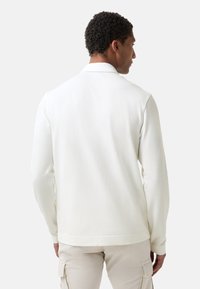 Mann mit kurzen lockigen Haaren, der ein weißes langärmeliges Hemd und beige Cargo-Hosen trägt, mit dem Rücken zu einer schlichten Hintergrund. U