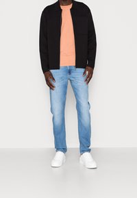 Svart zip-up jacka med bröstficka, persikofärgad crew-neck t-shirt, ljusblå jeans och vita sneakers med röda accenter.