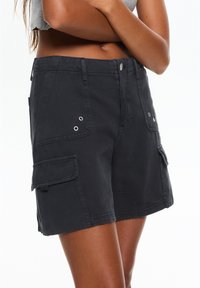 Pantaloni cargo neri realizzati in tessuto resistente, dotati di tasche frontali, chiusura con bottone e grommet decorativi. Lunghezza fino a metà coscia.