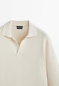 Kermanvärinen neulepolo, jossa on tekstuuri, v-pääntie ja perinteinen kaulus. Kankaasta näyttää olevan kevyt ja hengittävä.