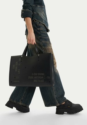 Osoba ubrana w ciemne dżinsowe ubrania i czarne, masywne buty, trzymająca dużą czarną torbę typu tote z nadrukiem tekstowym.