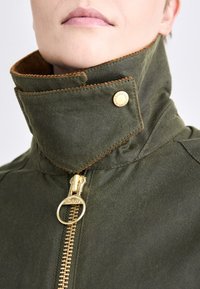 Olivově zelená voskovaná bunda s vysokým límcem, zlatým kováním a kontrastním prošíváním. Obsahuje logo na pullu zipu a accenty knoflíků.