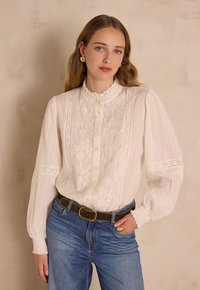 Camicia bianca con dettagli in pizzo, davanti con bottoni, colletto arricciato e maniche lunghe. Abbinata a jeans di denim blu e una cintura marrone.