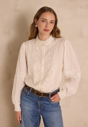Camicia bianca con dettagli in pizzo, davanti con bottoni, colletto arricciato e maniche lunghe. Abbinata a jeans di denim blu e una cintura marrone.