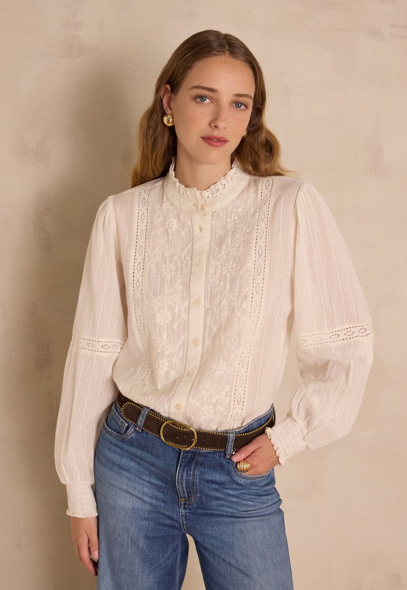 Camicia bianca con dettagli in pizzo, davanti con bottoni, colletto arricciato e maniche lunghe. Abbinata a jeans di denim blu e una cintura marrone.