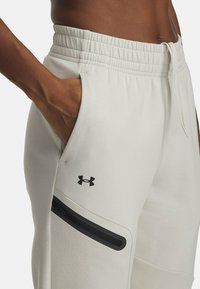 Light grey sweatpants with an elastic waistband, side pockets, and a zippered pocket. Features a black logo on the thigh and sleek design.