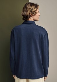 Marineblaue Langarmbluse aus strukturiertem Stoff, mit Kragen und lockerer Passform, und einem geraden Saum, der hinten sichtbar ist.