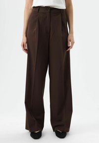 Pantalon marron à jambes larges en tissu lisse, avec une taille haute, des plis à l'avant et une fermeture par bouton sur le devant. Chaussures noires visibles.