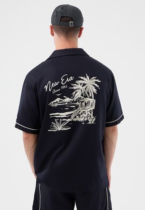 Vyras dėvintis juodą kepuraitę ir trumpomis rankovėmis marškinėlius su palmės medžio, gelbėtojo būdos ir užrašo „New Era Since 1920“ grafika ant nugaros.