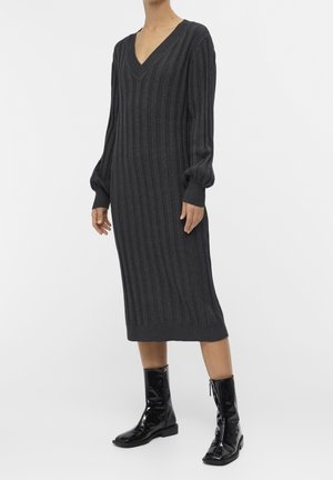 Femme portant une robe midi en maille côtelée gris foncé avec des manches longues bouffantes et des bottines noires brillantes, debout sur un fond blanc.