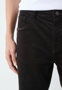 Svarte fløjlbukser med en struktureret overflade, der har forlommer, knaplukning i taljen og diskrete sømdetaljer.