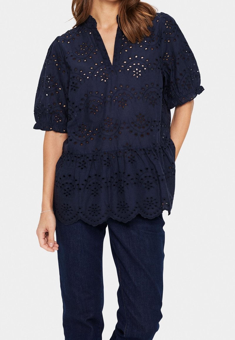 Femme portant un chemisier bleu marine à manches courtes avec motif ajouré et ourlet festonné, associé à un jean bleu foncé sur un fond uni.
