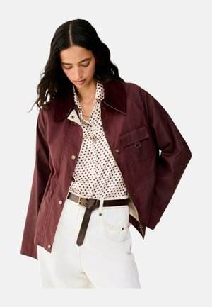 Femme portant une veste bordeaux sur une chemise blanche à pois, rentrée dans un pantalon blanc taille haute avec une ceinture noire, regardant vers le bas.