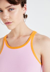 Lichtroze geribde tanktop met oranje bies, met een getailleerd ontwerp en brede schouderbanden. Bevat subtiele accenten van huidskleur.