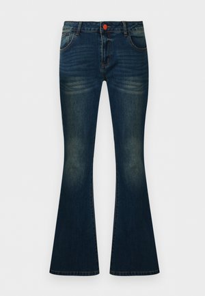 Femeie care poartă blugi evazați din denim albastru închis, top maro fără mâneci și sandale maro cu vârful deschis, stând în fața unui fundal simplu.