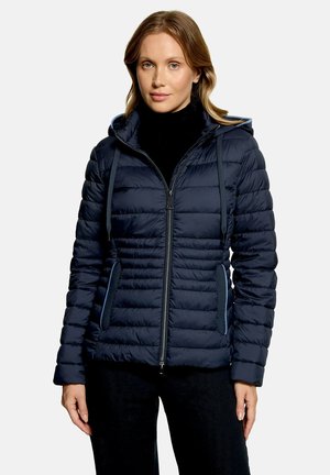 Femme portant une veste matelassée à capuche bleu marine avec fermeture éclair et poches latérales, debout devant un fond clair uni.